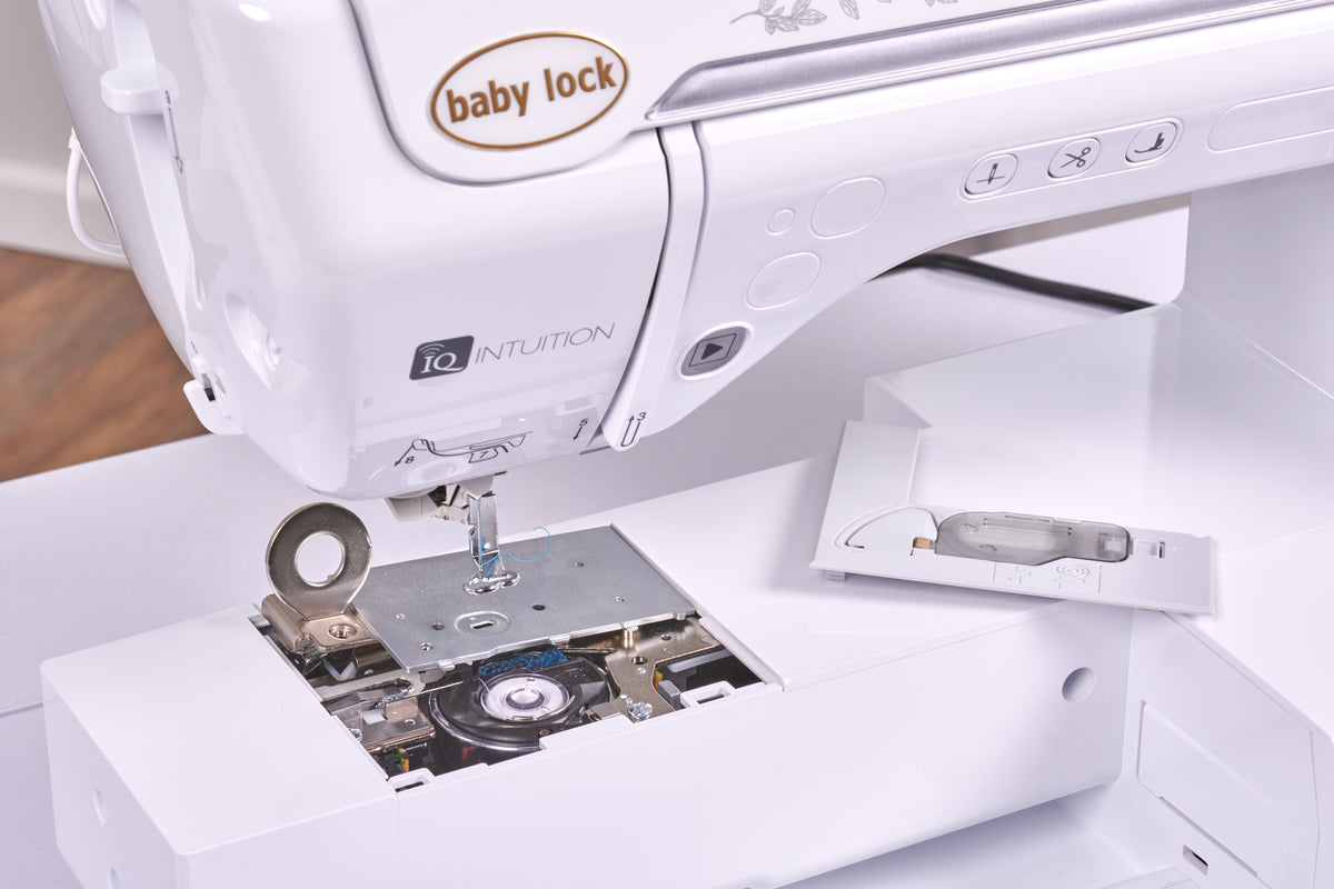 Close-up of a white Baby Lock Ursa Embroidery machine with the lower cover removed, showing internal components and a small instruction card beside the needle area.